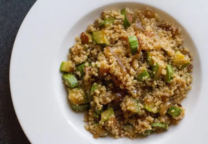 Un piatto di quinoa con verdure, tra cui zucchine a cubetti e cipolle caramellate, servito su un piatto bianco.