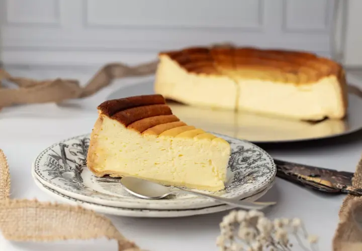 Une part de tarte au fromage, dorée et crémeuse, sur une assiette ornée. Le reste du gâteau est visible en arrière-plan.