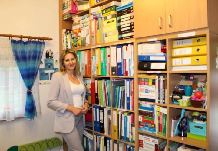 Kobieta w jasnej marynarce stoi w nowoczesnym gabinecie logopedycznym, otoczona regałami pełnymi materiałów terapeutycznych.