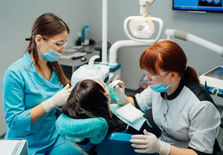 Kurs asystentka stomatologiczna: dwie uczące się kobiety w okularach i maseczkach pracują przy pacjentce w gabinecie dentystycznym.