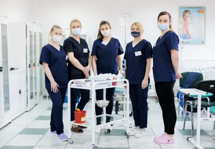 Grupa studentek pielęgniarstwa w maseczkach stoi przy wózku medycznym w sali szpitalnej.