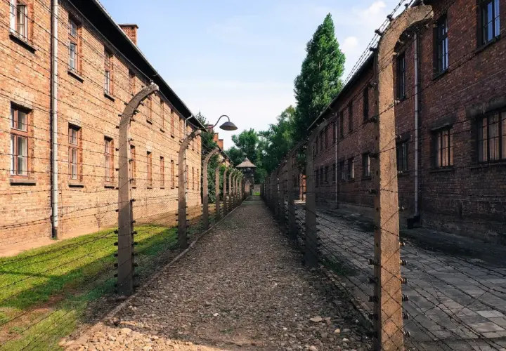 Ścieżka między ceglanymi budynkami, otoczona drutem kolczastym, prowadzi do wieży strażniczej. Miejsce pamięci Auschwitz Birkenau Museum.