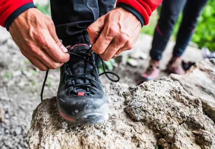 Dłonie zawiązują sznurówki czarnych butów podejściowych, gotowych na górską wyprawę. W tle widać inne buty trekkingowe.
