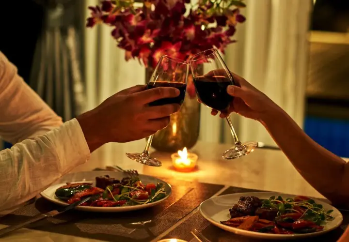 Romantyczna kolacja dla dwojga przy świecach. Dwie pary kieliszków z czerwonym winem unoszą się w geście toastu.