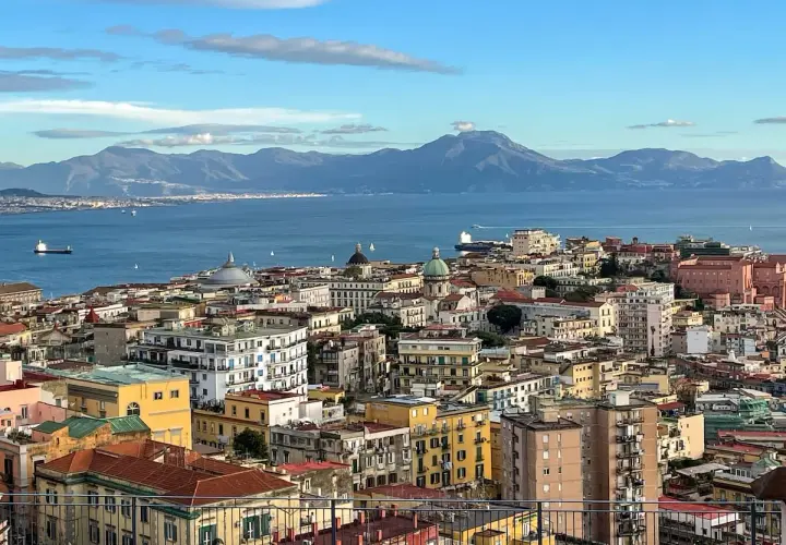 A panoramic view of Naples in December, with the blue sea and distant mountains under a cloudy sky.