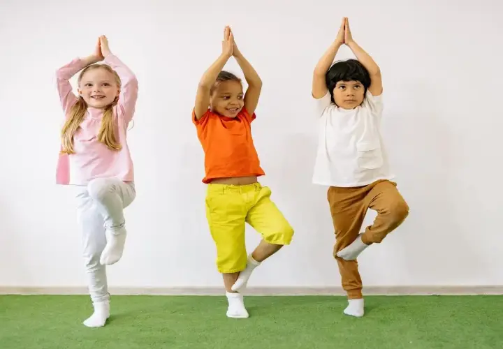 Tre bambini praticano yoga, in piedi su una gamba sola, con le mani giunte sopra la testa.