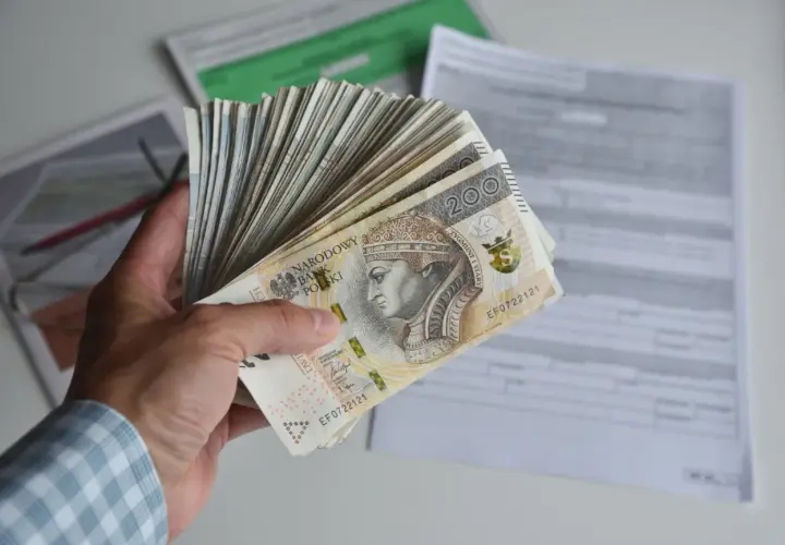 Dłoń trzyma plik banknotów 200 zł, symbolizujących dochód netto, na tle dokumentów podatkowych.