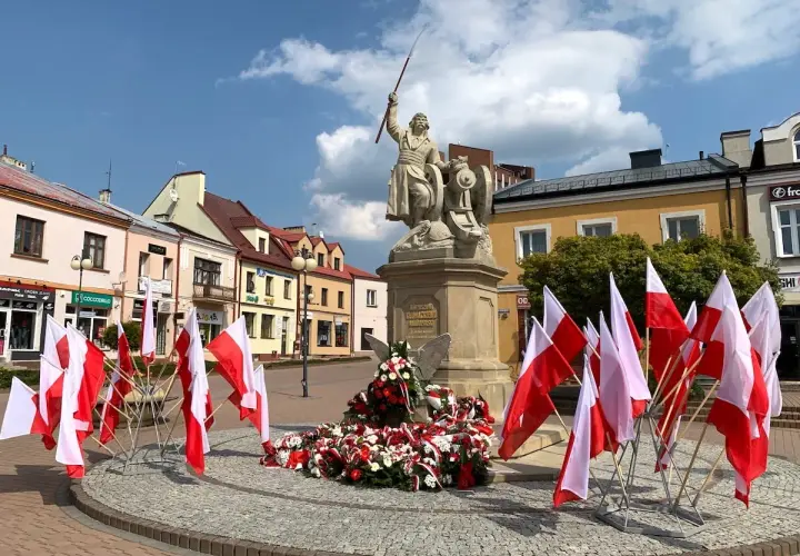 Pomnik z polskimi flagami, w tym kierunkowy 15, na placu przed kolorowymi kamienicami.