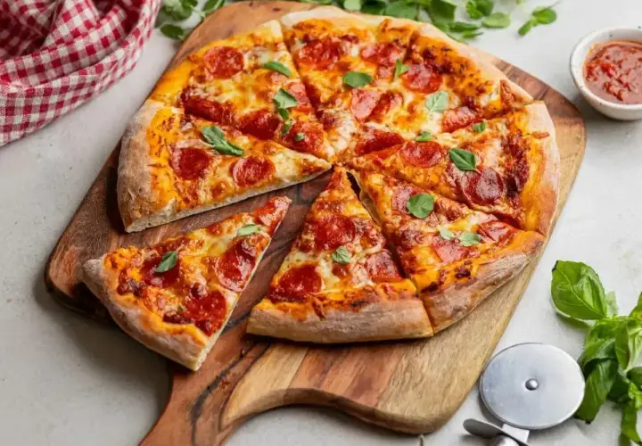 Pyszna pizza z pepperoni i świeżą bazylią, pokrojona na kawałki, leży na drewnianej desce. Obok sos pomidorowy i nóż do pizzy.