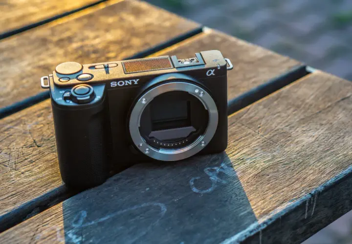 Czarny aparat Sony ZV-E10 II bez obiektywu leży na drewnianym stole, oświetlony ciepłym światłem zachodzącego słońca.