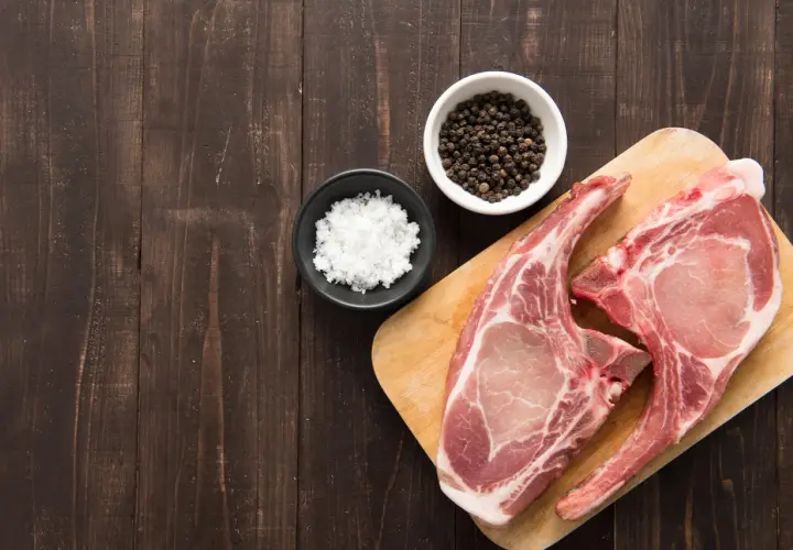 Deux belles côtes de porc crues sur une planche en bois, prêtes pour une cuisson au four, avec du sel et du poivre en grains à côté.