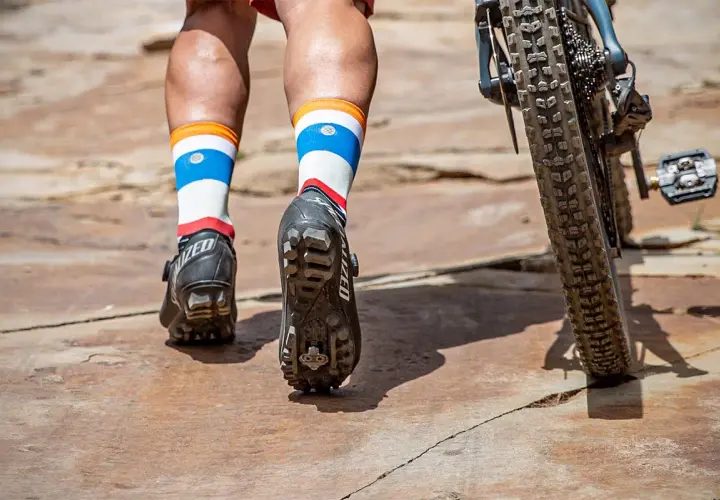 Mountain biking shoes with cleats are shown, not hiking boots. The rider's colorful socks and bike tire are also visible.