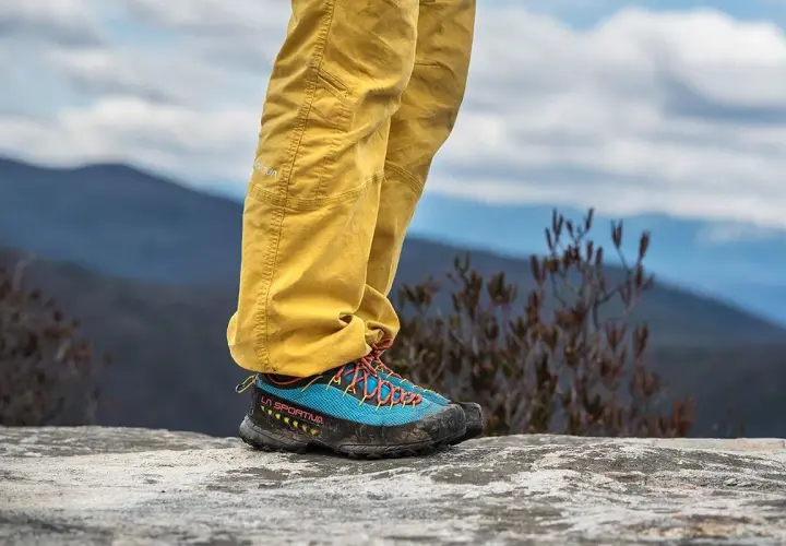A person stands on a rock, wearing yellow pants and blue approach shoes with orange laces, ready for an adventure.