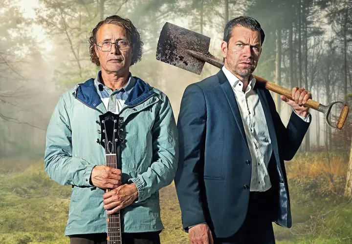 Två män i skogen, en med gitarr, en med spade. En scen från en mads mikkelsen serie.