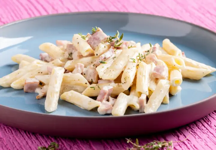 Penne pasta with creamy sauce and diced ham, garnished with thyme, on a blue plate.
