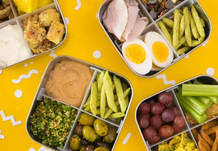 Four bento boxes filled with colorful, healthy foods, perfect for bento for beginners. Includes chicken, eggs, olives, grapes, and more.