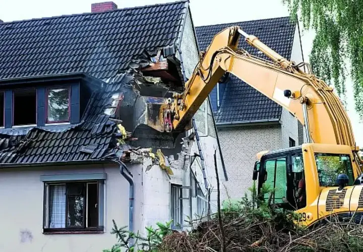 Żółta koparka prowadzi rozbiórkę dachu domu, niszcząc jego konstrukcję.