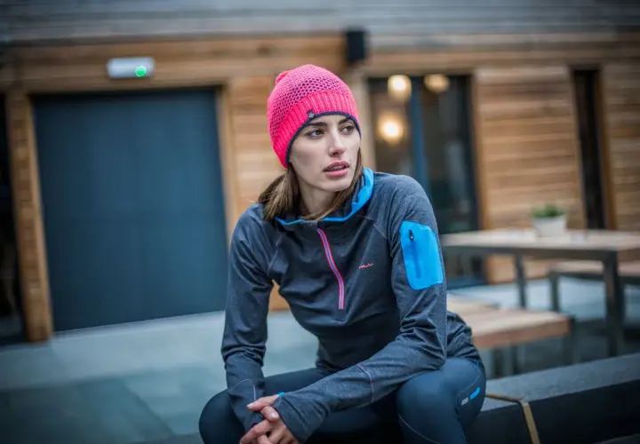 Kobieta w sportowym stroju, z jaskrawym, różowym czapka beanie, siedzi na ławce przed nowoczesnym budynkiem.