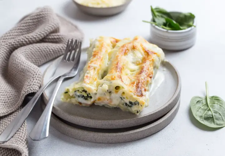 Pyszne cannelloni ze szpinakiem, zapieczone pod beszamelem i serem, na talerzu z widelcem i serwetką.
