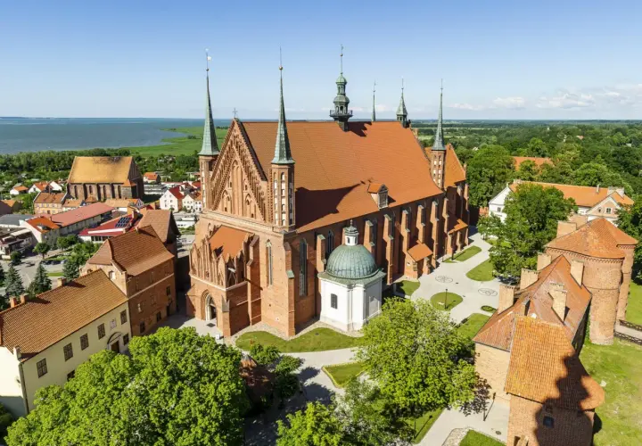 Zamek Frombork z lotu ptaka, z dominującą bazyliką katedralną i wieżami obronnymi, nad błękitną wodą Zalewu Wiślanego.