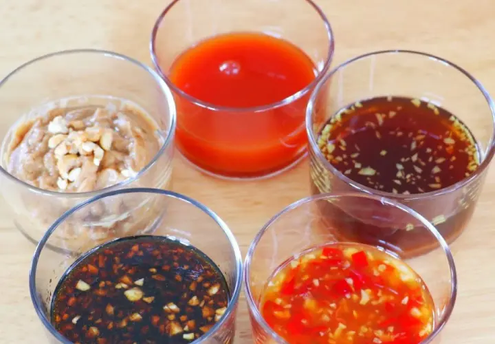 Five small glasses hold various Japanese dipping sauces, including a creamy peanut sauce, a bright red sauce, and a dark soy-based sauce with garlic.