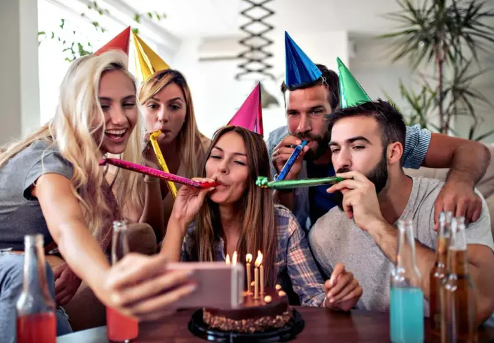 Grupa przyjaciół świętuje 40 urodziny w domu, dmuchając w trąbki i robiąc selfie z tortem.
