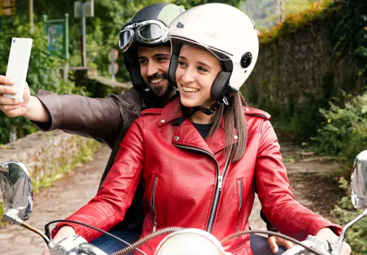 Pareja en moto, él con casco y gafas, ella con casco blanco y chaqueta roja, toman una selfie con el móvil. ¡Manos libres, casco moto legal y a disfrutar del viaje!