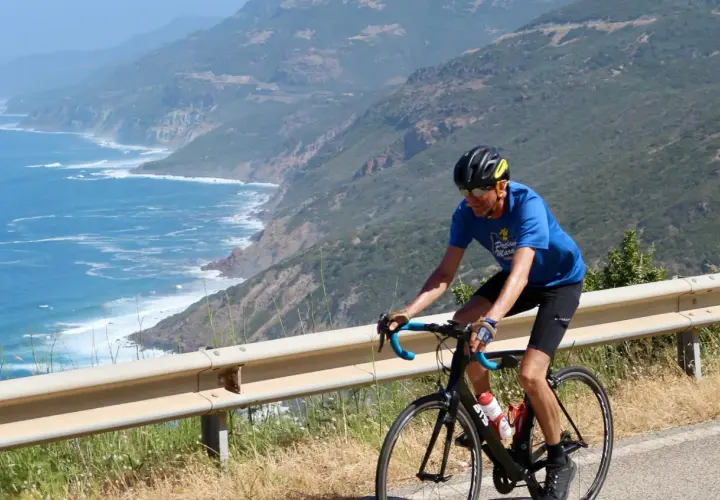 Man on a bike tour in Sardinia, cycling along a scenic coastal road with the blue sea and green hills.