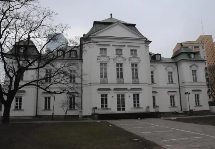 Pałac Przebendowskich, biała, elegancka budowla z zielonym dachem, otoczona drzewami i nowoczesnymi budynkami.