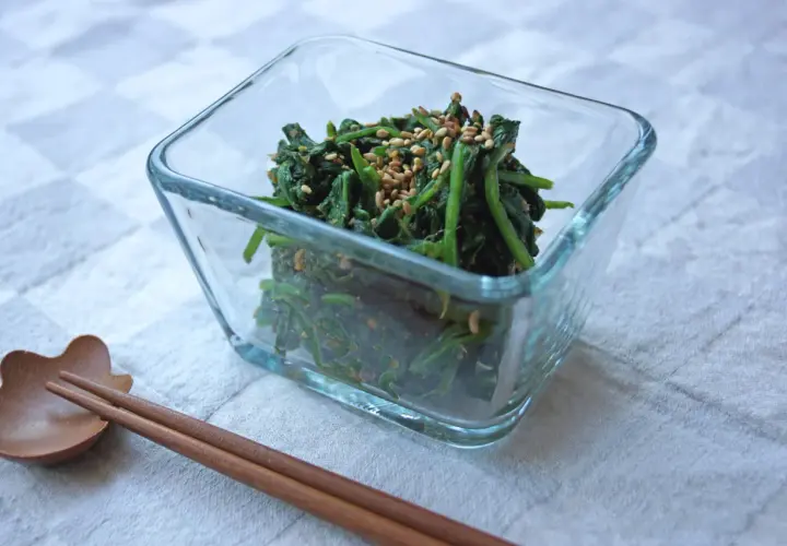 A glass bowl filled with spinach gomae, a savory sesame-dressed spinach dish, is ready to be enjoyed with chopsticks.