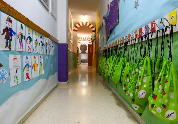 Pasillo de guardería en Comunidad Valenciana. Dibujos infantiles decoran la pared, y mochilas verdes cuelgan listas para la ayuda.