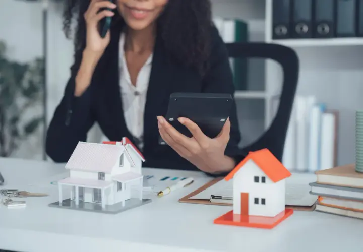 Agent immobilier au téléphone, calculatrice en main, devant des maquettes de maisons. Elle prépare une vente à terme.