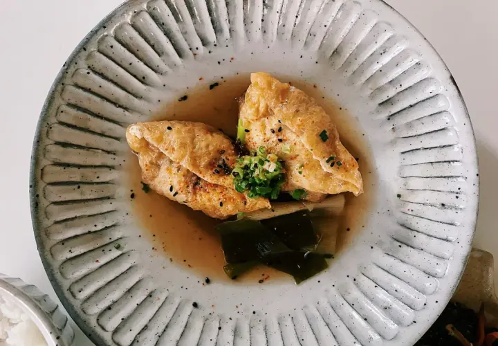 Two pieces of golden aburaage tofu simmer in a savory broth with leeks and seaweed, garnished with green onions and sesame seeds.