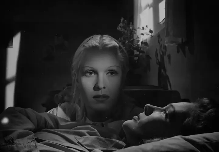 De två kvinnorna i dramatisk belysning. En kvinna ligger ner, medan den andra tittar ner på henne med intensiv blick.
