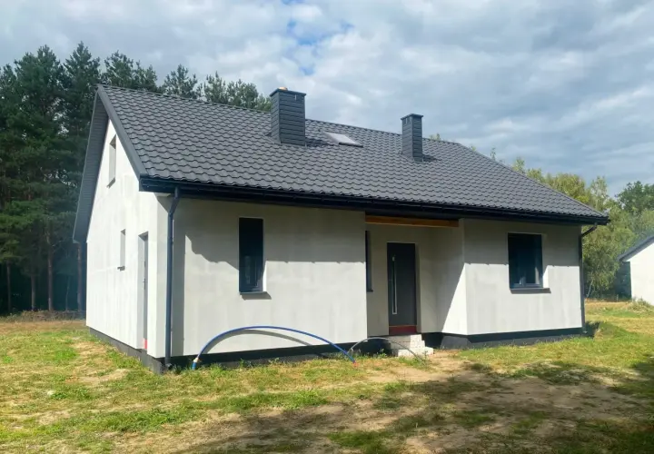 Nowy dom szkieletowy na lata, z szarym dachem i białymi ścianami, otoczony zielenią i lasem.