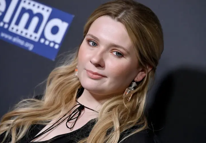 Abigail Breslin, znana z wielu filmów, z niebieskimi oczami i blond włosami, pozuje z uśmiechem.
