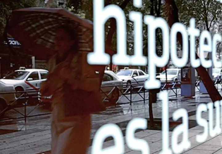 Reflejo en un escaparate: "Hipoteca, ¿es la fein suya?". Una persona con paraguas pasa por la calle lluviosa con taxis.
