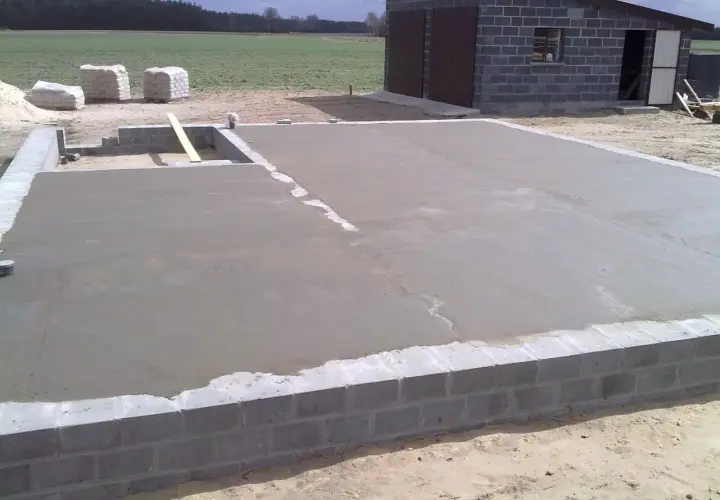 Świeżo wylana płyta betonowa, z widoczną minimalną grubością chudziaka, na fundamencie z bloczków. W tle budynek gospodarczy.