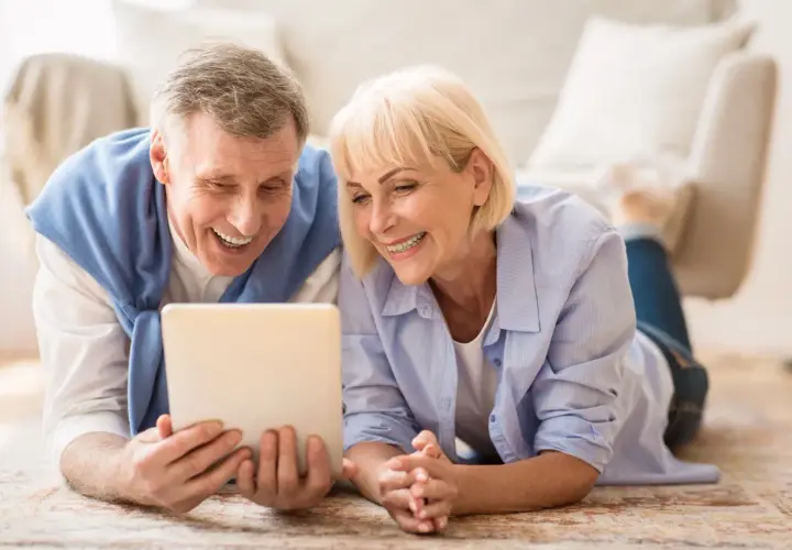 Ein lachendes älteres Paar liegt auf dem Boden und schaut auf ein Tablet. Ihre positiven senioren dates erfahrungen werden durch ihr Lächeln deutlich.