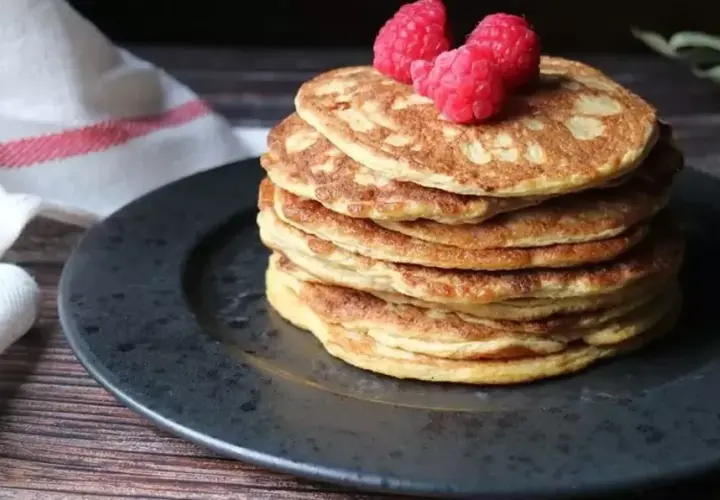 En hög med nyttiga pannkakor toppas med hallon, serverade på en mörk tallrik med en vit handduk med röda ränder i bakgrunden.