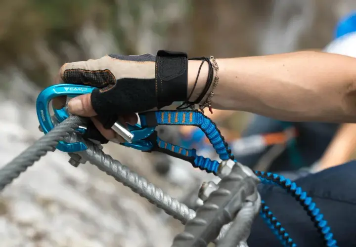 Ręka w rękawiczce z karabinkiem i lonżą, kluczowy element via ferrata sprzętu, zabezpiecza wspinaczkę.