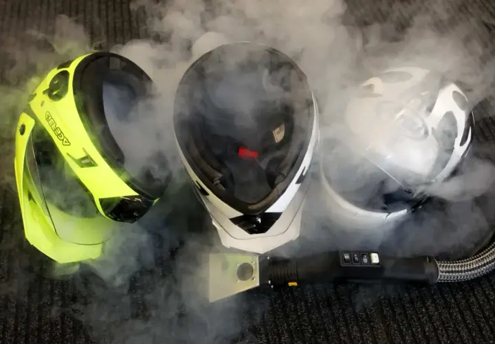 Tres cascos de moto rodeados de vapor. Uno amarillo brillante, uno blanco y negro, y uno blanco con diseño. Un limpiador a vapor está cerca. ¿Son estas las peores marcas de cascos para moto?