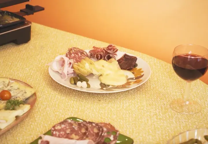 Un verre de vin rouge accompagne une raclette savoureuse, des charcuteries et des fromages sur une table jaune.