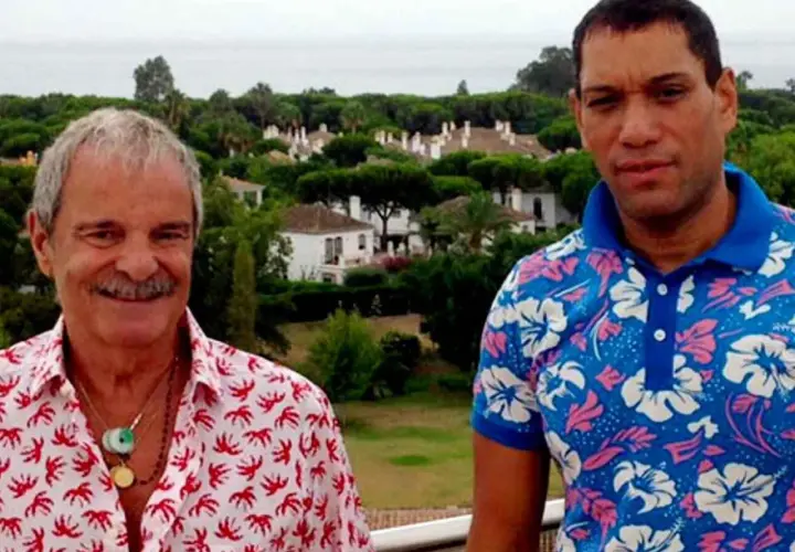 Elio Valderrama Prescott y un amigo posan con camisas coloridas frente a un paisaje de casas y árboles.