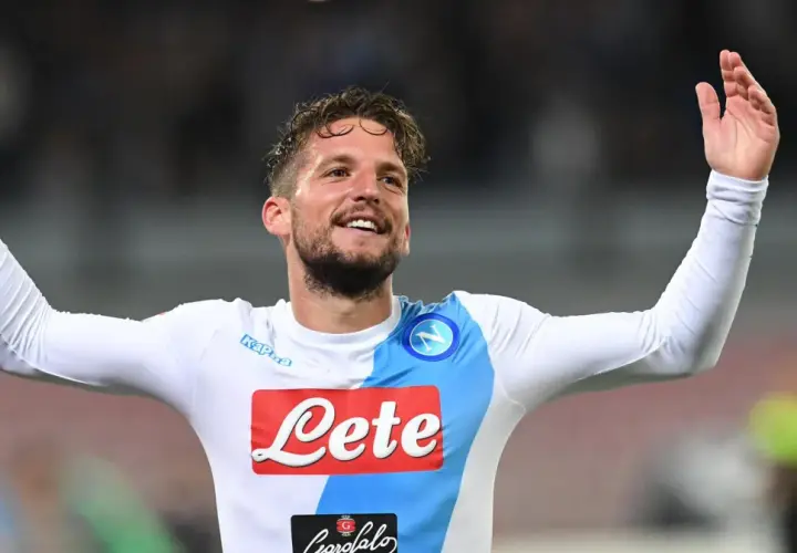 Uśmiechnięty Dries Mertens w białej koszulce Napoli z rękami uniesionymi w geście radości.