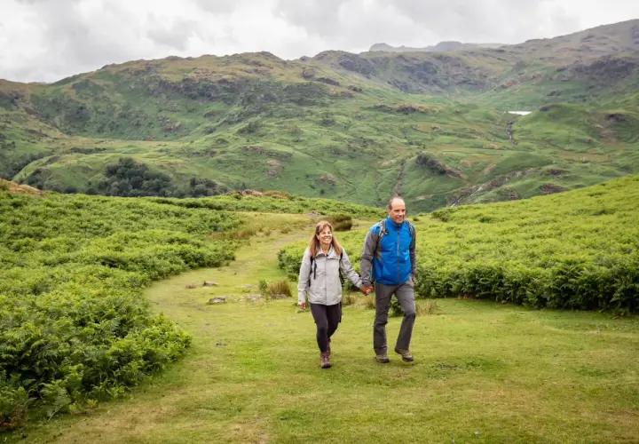 A couple enjoys hiking in England, hand-in-hand, amidst lush green hills and ferns under a cloudy sky.
