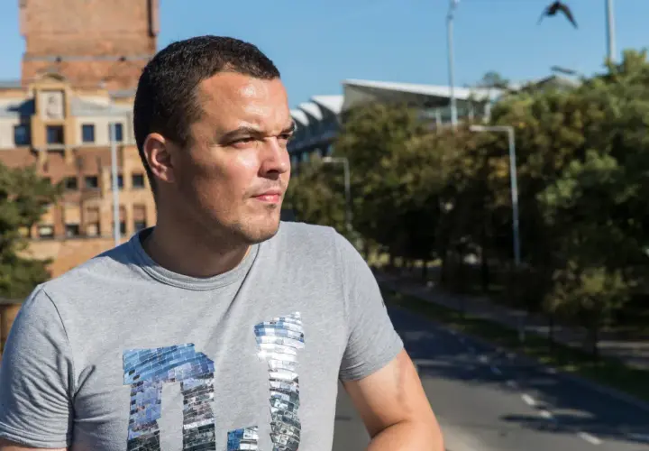 Dawid Janczyk w szarej koszulce z błyszczącym napisem, na tle miejskiego krajobrazu z budynkami i drzewami.