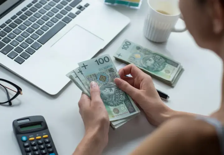 Dłonie trzymające polskie banknoty 100 zł obok laptopa, okularów i kalkulatora.