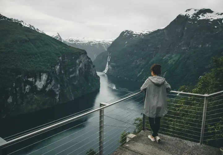 Kobieta podziwia majestatyczne fiordy norweskie, z wodospadem w oddali i ośnieżonymi szczytami gór.