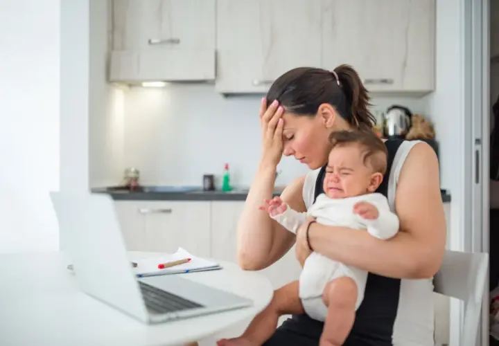 Zmęczona mama z płaczącym, **high need baby** na rękach, próbuje pracować przy laptopie.